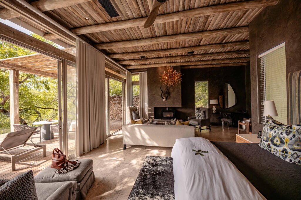 Kapama Karula villa bedroom opening onto a shaded deck, with a lounge and fireplace beyond