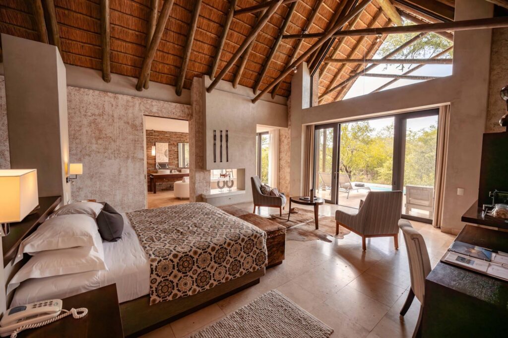 Kapama Southern Camp Luxury Villa bedroom under a thatched roof with glass doors to the bush